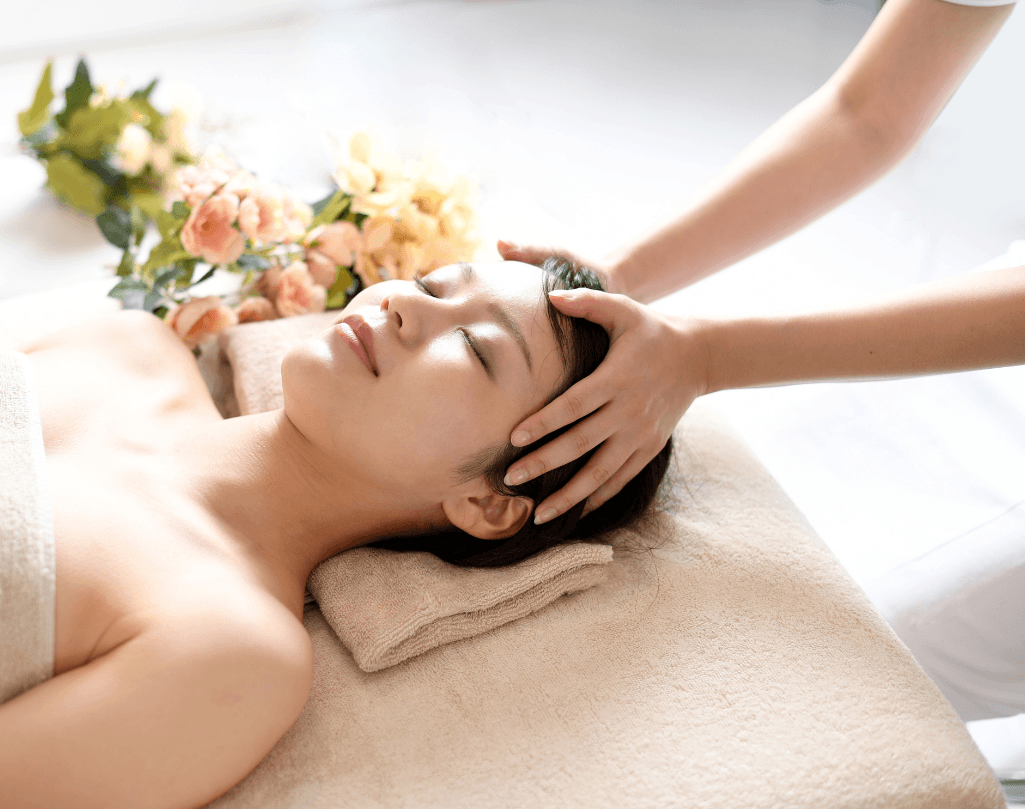 Woman lying peacefully on a spa bed receiving a relaxing head massage, with soft lighting and floral decoration in the background