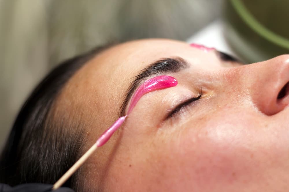 Close-up of a woman receiving a professional eyebrow wax, with a strip placed above the brow and gloved hands preparing the area, highlighting precise facial hair removal