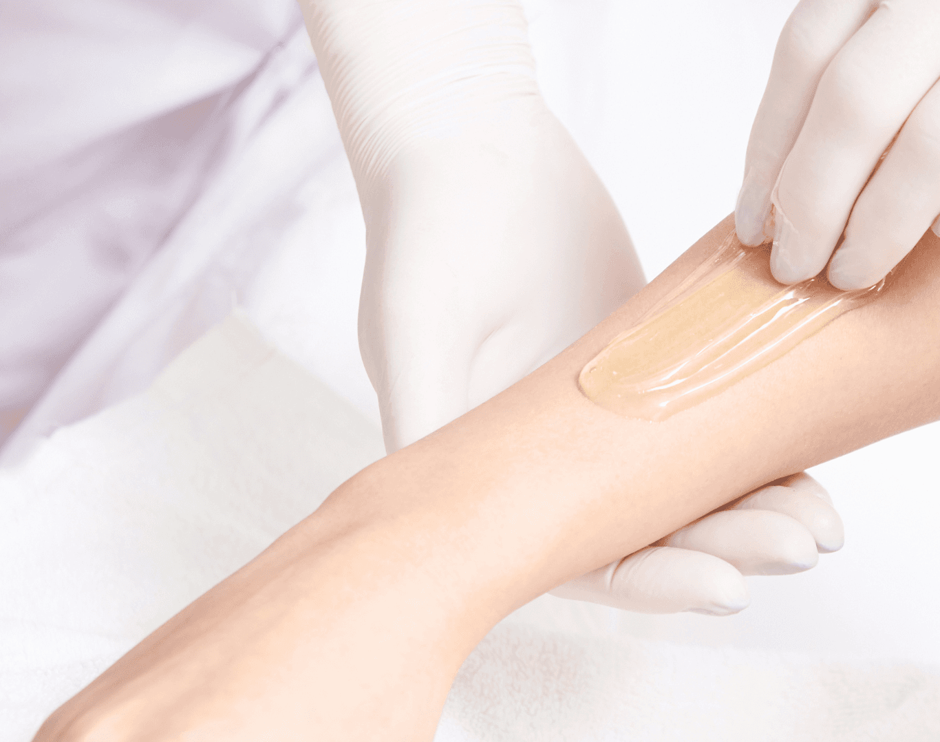 A close-up of a therapist applying warm wax to a client’s forearm in preparation for hair removal, using a gloved hand for precision and hygiene