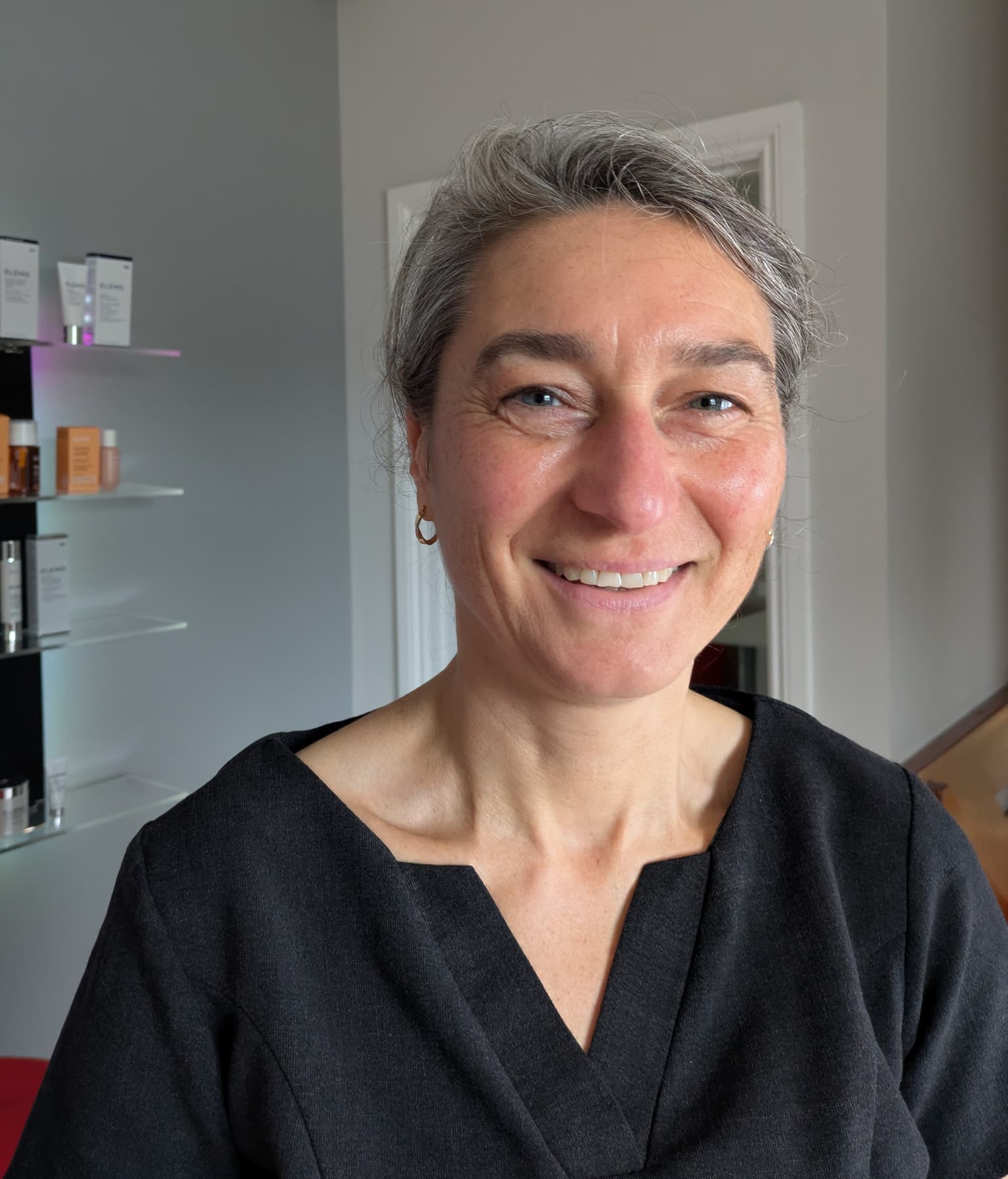 Woman with grey hair tied back, wearing a black uniform top, smiling indoors with skincare products displayed on shelves in the background.