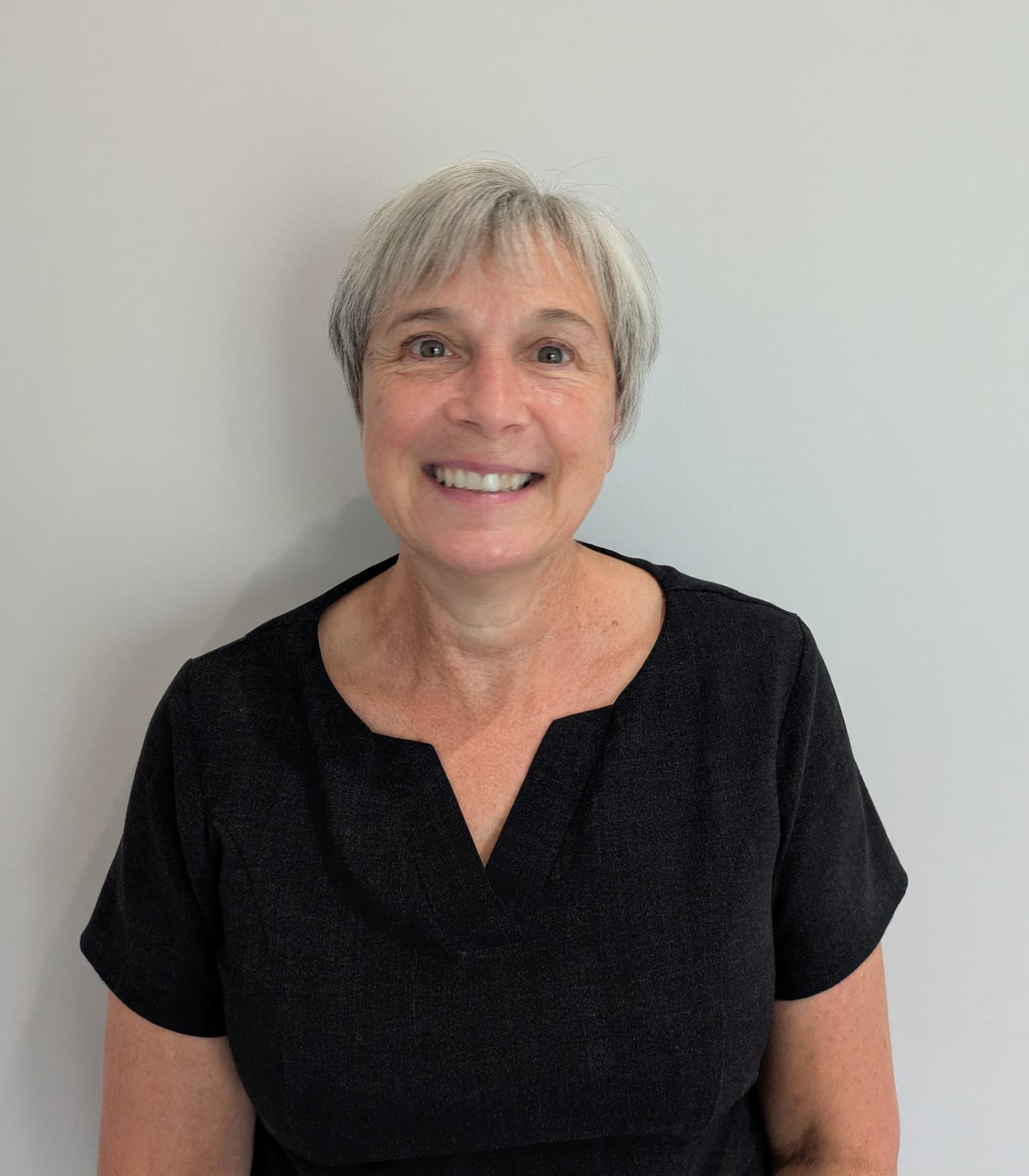 Woman with short grey hair wearing a black uniform top, smiling against a plain light-coloured background.