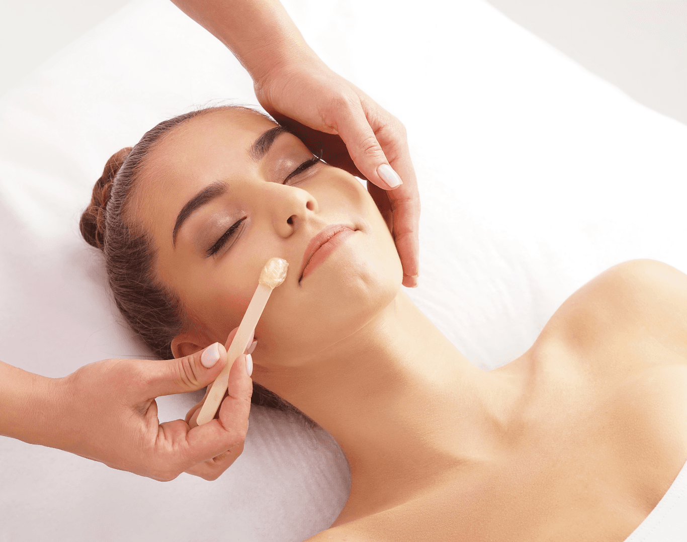 Woman lying on a treatment bed with eyes closed while a beauty therapist applies wax to her upper lip, representing a calm and professional waxing experience.
